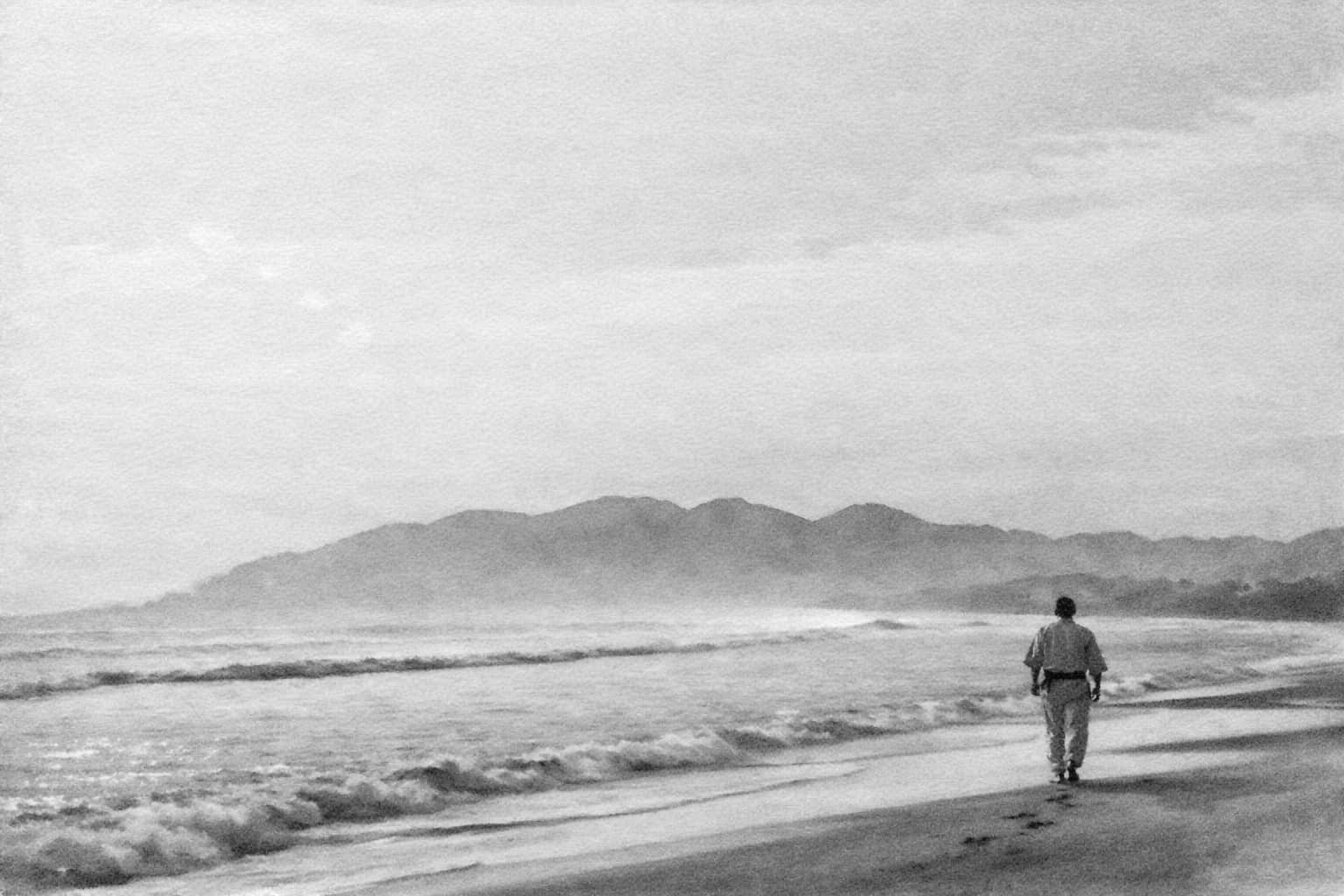 Sosai Masutatsu Oyama walking alone by the sea in soft dawn light. The open horizon and distant mountains reinforce the impression of solitary training, reflection, and a lifelong search for truth through budō.