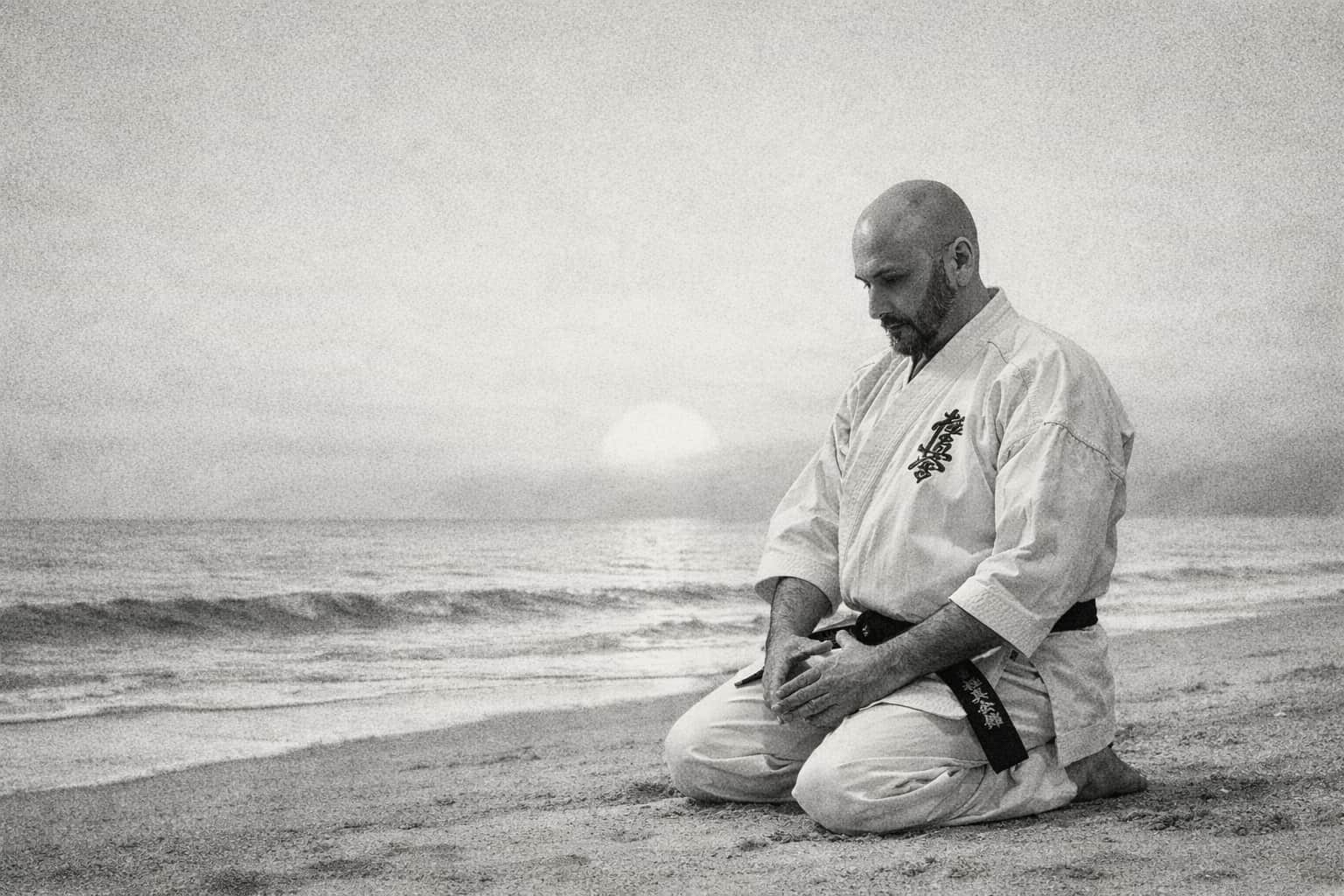 Black and white image of a man in a karate gi sitting in seiza on a beach by the sea.